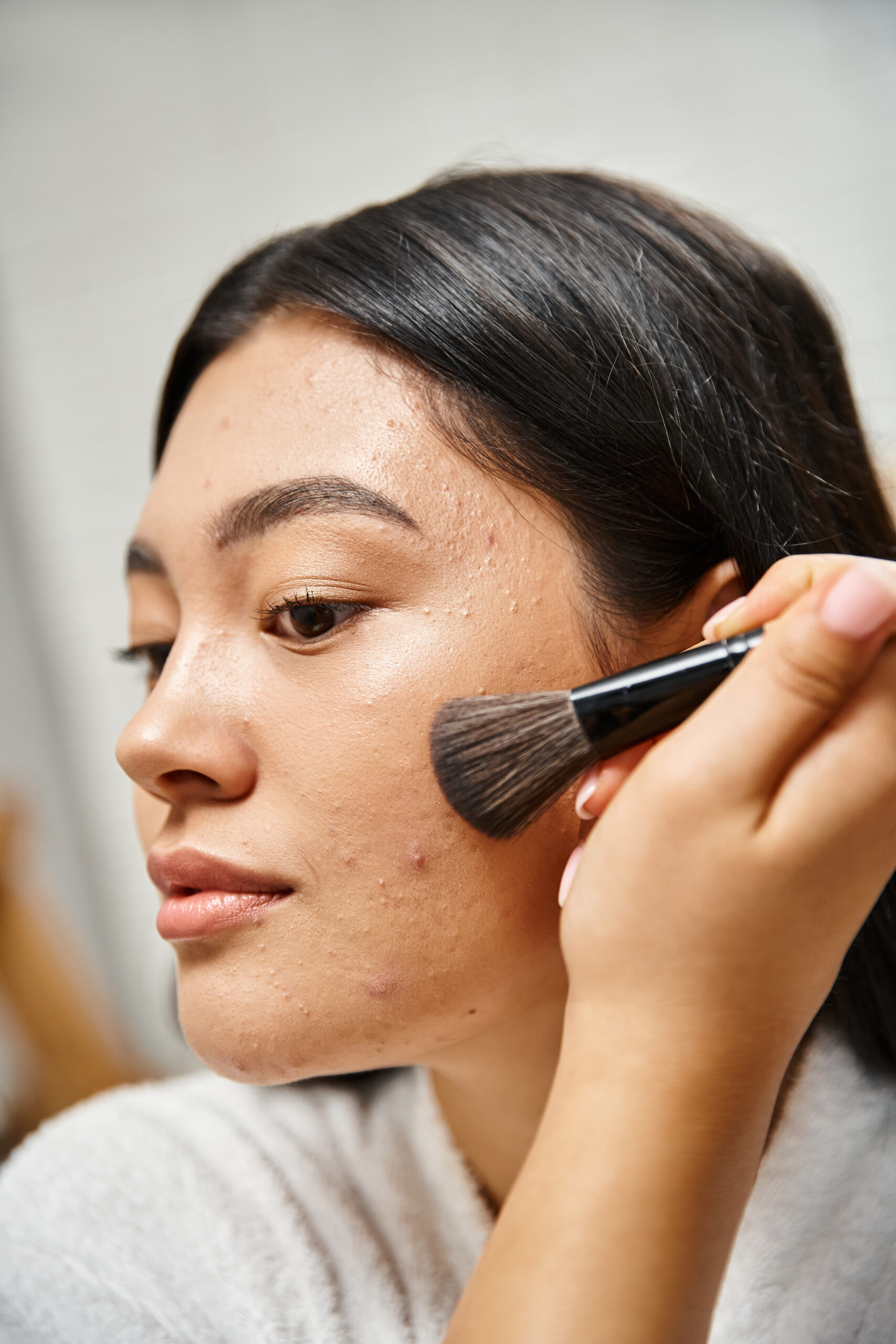 brunette and young asian woman applying face powder over acne prone skin with cosmetic brush
